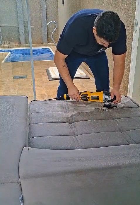 Profissional da JV Clean de uniforme azul-marinho trabalhando com equipamento no assento de um sofá cinza.